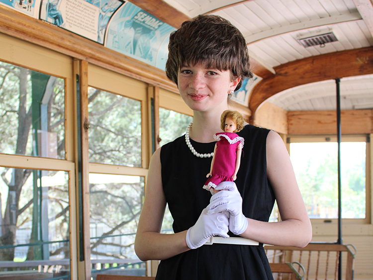 A young girl dressed in a black dress, white gloves and a pearl necklace holds a vintage Barbie doll while standing inside a historic train car.