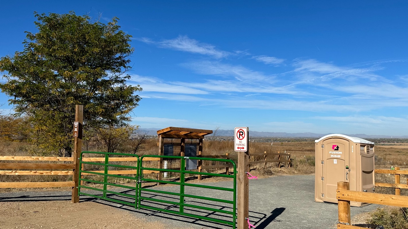 A dirt trailhead with a tan portable restroom and green gate with a grassy prairie in the background