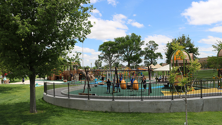 Green grass, trees, and a black metal fence surround a yellow, green, and blue playground with accessible swings and ramps.