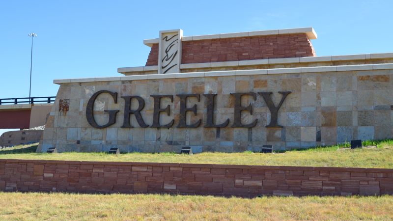 A large stone and brick welcome sign displaying "GREELEY" in bold, metal lettering, set against a clear blue sky at a highway overpass.