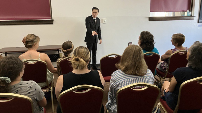 A young person wearing a black suit presents to a crowd of people seated in the community room at the Greeley History Museum.