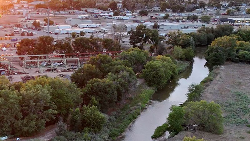 Aerial view of the Poudre River in Greeley, bordered by dense trees with industrial buildings and warehouses on the left with a small urban area in the background. An orange sunset casts a warm glow over the landscape.