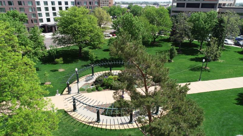 An aerial view of a curved walkway with decorative metal archways in an urban park setting.  The walkway is surrounded by well-maintained green lawn and mature trees.