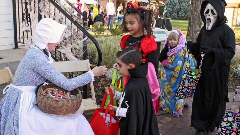 Children dressed in costumes hold bags out for person dressed in costume to fill with candy as she stands on porch of historical building.