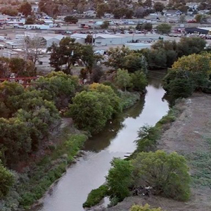 Aerial view of the Poudre River in Greeley, bordered by dense trees with industrial buildings and warehouses on the left with a small urban area in the background. An orange sunset casts a warm glow over the landscape.