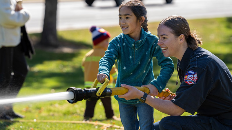 Female firefighter helping a young girl use a fire hose at an event, with a young child dressed in a firefighter outfit standing in the background.