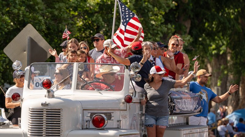 Men, women and children smiling, waving, and carrying American flags while riding on an old-fashioned truck