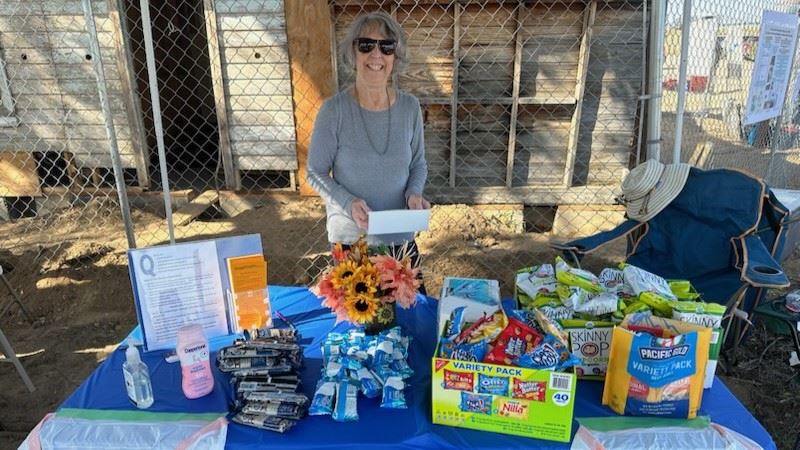 Susan Cribelli stands behind a table covered with snacks, a bouquet of flowers and trash bags at the Dearfield Day event. The former Dearfield filling station is in the background.