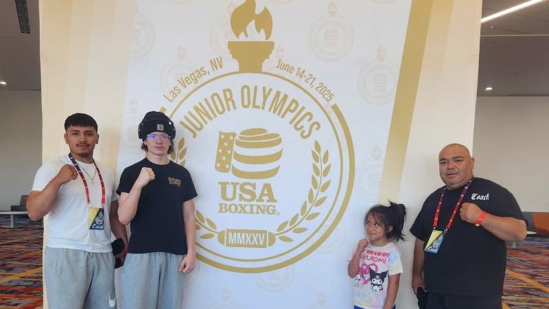 Two boxers stand with their boxing coaches in front of the Junior Olympics banner at the Las Vegas Convention Center.